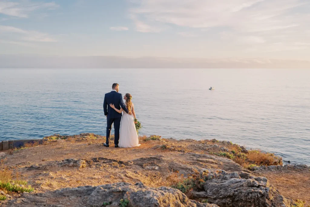 Couple, Bride and Groom at the clifs of Cascais at the end of the day with warm light, magnificent sea view, near the Guia Lighthouse on their wedding day in Cascais.