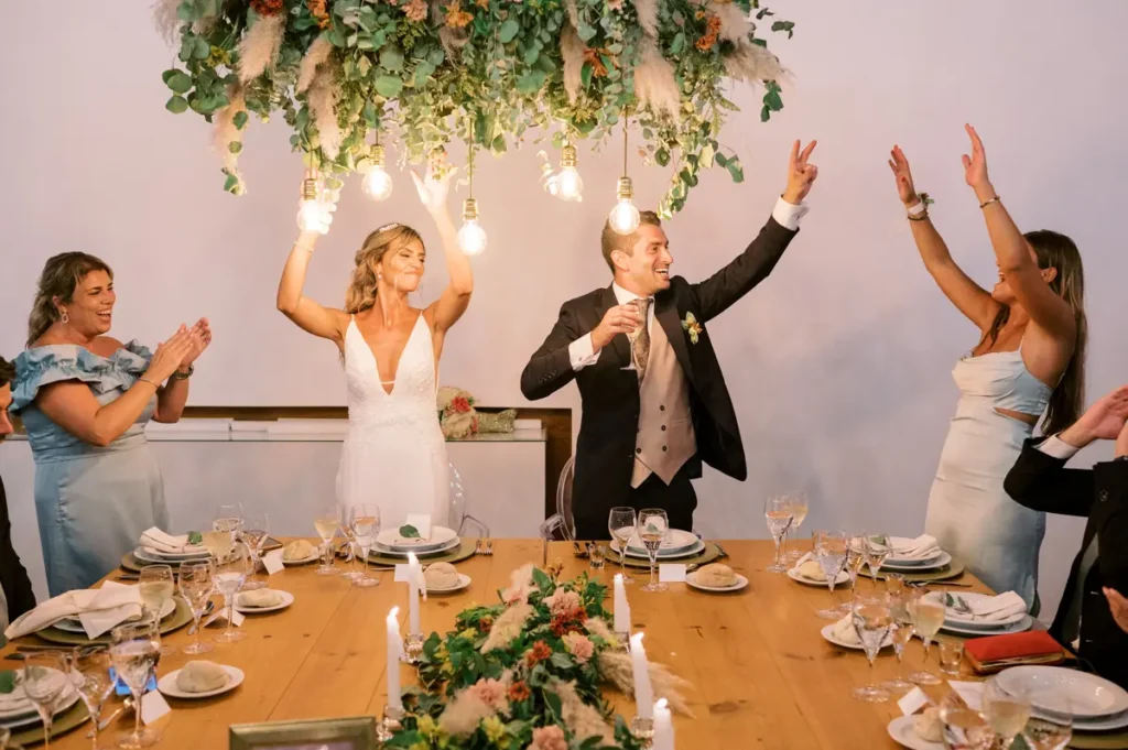 Noivos celebram com os convidados na mesa presidencial da Quinta da Bichinha, sob uma instalação floral suspensa com luzes Edison, captando a atmosfera festiva e elegante do jantar.