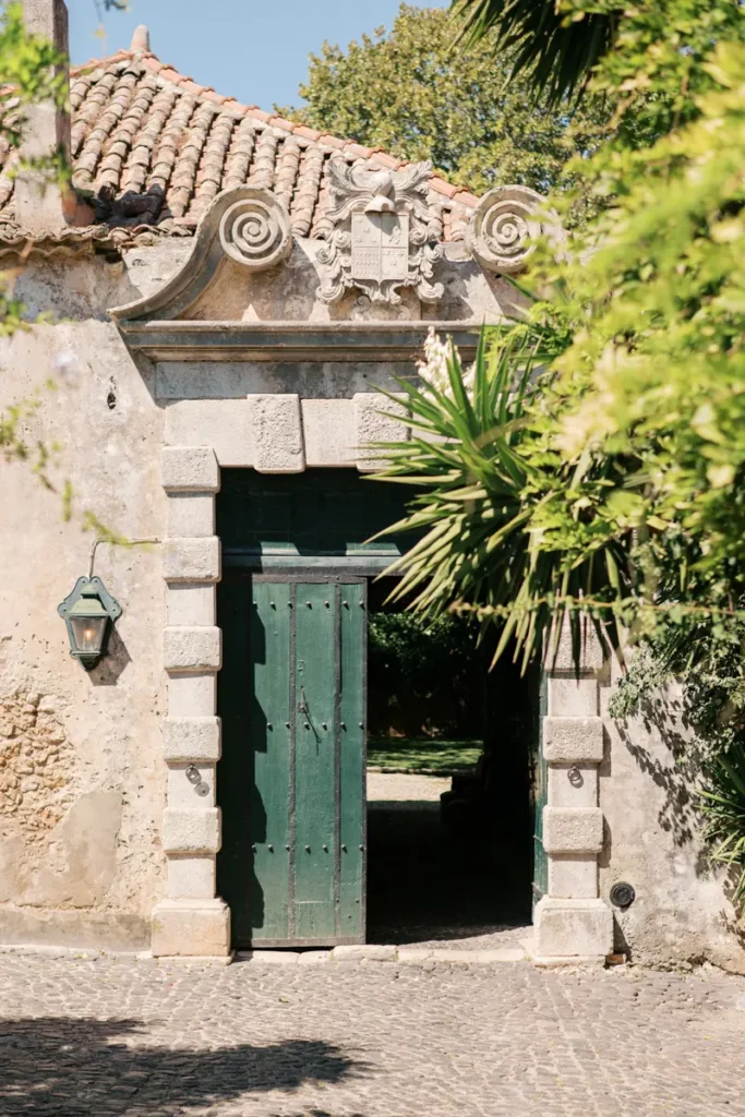 The imposing green wooden entrance door of Quinta do Torneiro, framed by classic stonework and topped with a historic coat of arms. This rustic 18th-century facade, flanked by lush foliage, represents the authentic Portuguese architecture that welcomes guests to your destination wedding near Lisbon.