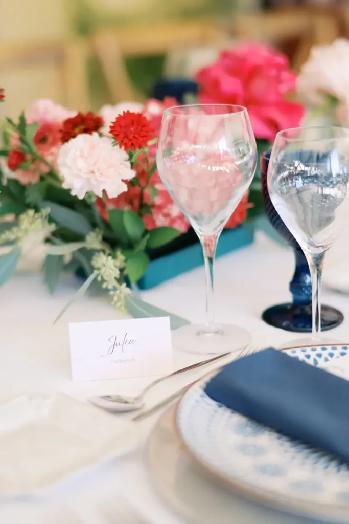 Elegant table decor detail for a wedding at Quinta do Torneiro, featuring a calligraphy place card for 'Julia Carranza', crystal glassware, and a textured cobalt blue water goblet. A vibrant floral arrangement in shades of pink and red with eucalyptus foliage adds color, while patterned tableware with a blue napkin invokes Portuguese tradition, exemplifying the ideal modern elegance for a destination wedding near Lisbon.