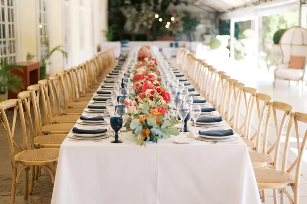 A long, elegantly decorated banquet table at Quinta do Torneiro, Oeiras, Portugal, stretching into perspective. The long table, draped with a white tablecloth, is lined on both sides by rustic wooden and cane chairs. Down the center runs a lush, colorful continuous floral runner made of fresh flowers in vibrant shades of pink, red, and orange (peonies, ranunculus) mixed with eucalyptus. The individual place settings feature white plates, navy blue linen napkins, and multiple clear and blue wine and water glasses. The semi-covered space features a historic wall with windows on the left and fairy lights hanging in the distance beneath the ceiling structure, evoking a luxurious and authentic celebration.