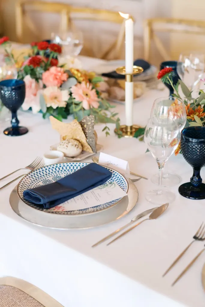 Elegant table decor detail for a wedding at Quinta do Torneiro, featuring a calligraphy place card for 'Julia Carranza', crystal glassware, and a textured cobalt blue water goblet. A vibrant floral arrangement in shades of pink and red with eucalyptus foliage adds color, while patterned tableware with a blue napkin invokes Portuguese tradition, exemplifying the ideal modern elegance for a destination wedding near Lisbon.