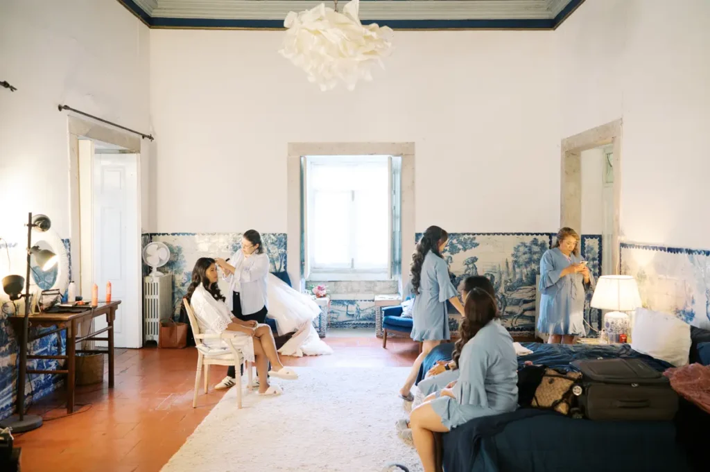 A candid documentary photo of a bride and her bridesmaids getting ready for a wedding in a historic master suite in Portugal. The bride, in a white robe, is sitting on a chair getting her hair done by a professional. Several bridesmaids in blue denim-style dresses are in the room; one is sitting on the edge of a large bed with a blue bedspread, and another is on the phone. The spacious room features white walls, terracotta flooring, and wainscoting covered with traditional blue and white scenic Portuguese tiles. There is a large window in the background, luggage, and a makeup table.