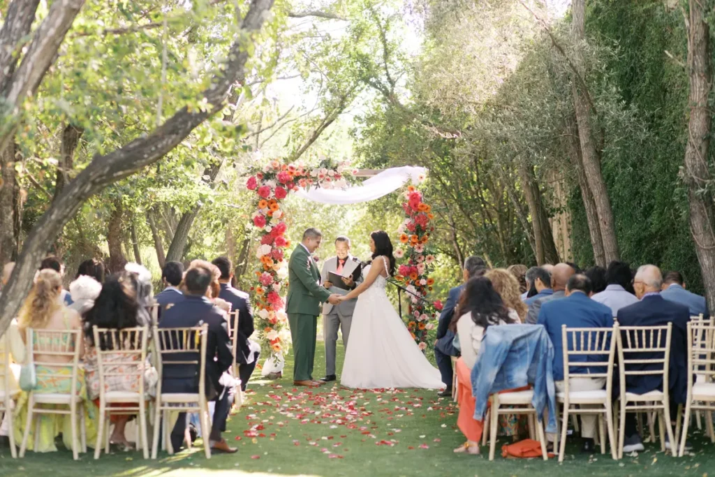 An outdoor wedding ceremony in a garden at Quinta do Torneiro. A couple, the groom in a green suit and the bride in a white dress, stand under a lush floral arch of pink, orange, and red flowers with white draping, exchanging vows before an officiant. Guests are seated on gold Chiavari chairs lined up on artificial grass beneath a canopy of leafy trees. Flower petals cover the center aisle. The background features a stone wall covered in greenery. Sunlight shines through the leaves.