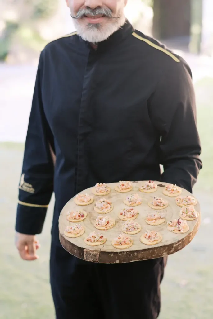 Detail of the premium catering service at a wedding at Quinta do Torneiro. A friendly waiter, dressed in an elegant black uniform with gold details, serves exquisite canapés and appetizers on a rustic wooden board during the outdoor cocktail hour. An excellent showcase of the gastronomic quality and attentive service for your destination wedding in Portugal.