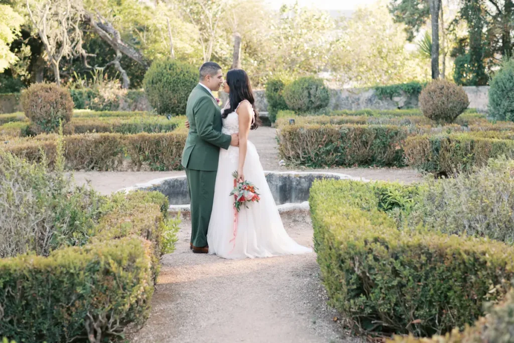 Um casal de noivos posa romanticamente num jardim formal e histórico na Quinta do Torneiro. O noivo, em fato verde-azeitona, e a noiva, em vestido branco segurando um bouquet floral, estão abraçados e prestes a beijar-se, emoldurados por sebes de buxo podadas e caminhos de cascalho, sob a luz suave de final de tarde. Uma fonte de pedra está visível ao fundo.