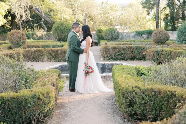 Um casal de noivos posa romanticamente num jardim formal e histórico na Quinta do Torneiro. O noivo, em fato verde-azeitona, e a noiva, em vestido branco segurando um bouquet floral, estão abraçados e prestes a beijar-se, emoldurados por sebes de buxo podadas e caminhos de cascalho, sob a luz suave de final de tarde. Uma fonte de pedra está visível ao fundo.