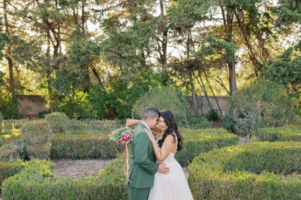 A bride and groom pose romantically in a formal, historic garden at Quinta do Torneiro. The groom, in an olive-green suit, and the bride, in a white dress holding a floral bouquet, are embracing and about to kiss, framed by manicured boxwood hedges and gravel paths under the soft late afternoon light. A stone fountain is visible in the background.