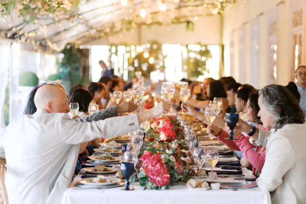 The same long table at Quinta do Torneiro from the previous image, now filled with wedding guests raising their glasses for a toast during the reception meal. The detailed floral runner with peonies and ranunculus and the hanging fairy lights are present, but now the table is full of life with guests celebrating.