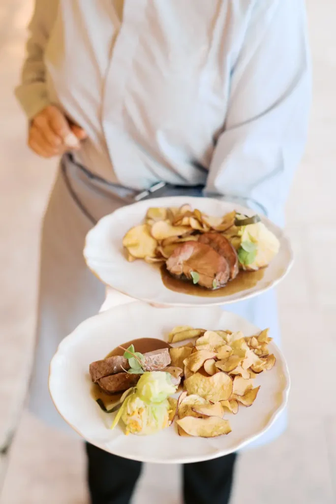 Exquisite detail of the main meat course plating at a wedding at Quinta do Torneiro. A catering staff member holds two classic plates featuring slices of tender meat drizzled with sauce, accompanied by crispy potato chips and an elegant bundle of fresh vegetables. An excellent representation of the gastronomic quality and service excellence available for your destination wedding in Portugal.