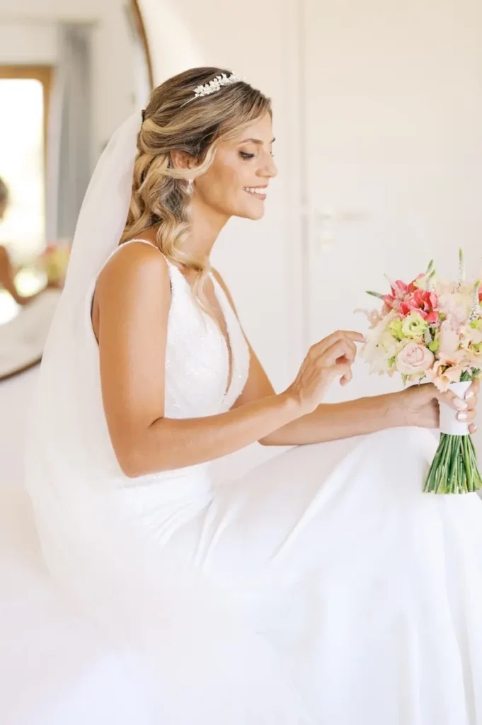 Side portrait of a smiling bride holding her flower bouquet at Quinta da Bichinha, showing off the veil, tiara, and dress details in natural light.