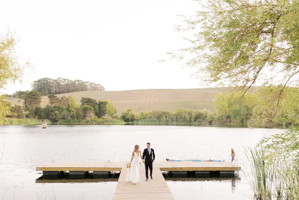 Bride and groom walking hand in hand along the lake pier at Quinta da Bichinha, with a panoramic view of the vineyards and surrounding nature in Alenquer. Photo captured by Boris The Cat - Wedding Photography.