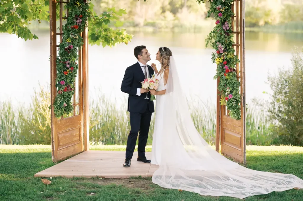 Bride and groom under a floral arch by the lake at Quinta da Bichinha, highlighting the elegance of the long veil and the natural setting of Alenquer for an outdoor ceremony.