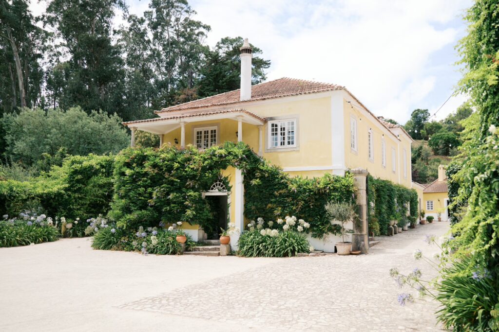 Vista exterior da casa principal da Quinta Nova do Hespanhol, um local de eventos em Portugal. O edifício de dois andares com paredes amarelas e varanda de colunas é coberto por trepadeiras com flores roxas, ladeado por agapantos na base. 
