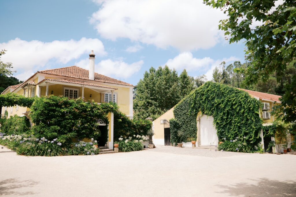 Visão externa externa da Quinta Nova do Hespanhol, um local de eventos em Portugal, sob um céu azul nublado. O edifício principal de dois andares com paredes amarelas e varanda de colunas é coberto por trepadeiras com flores roxas, ladeado por agapantos na base. Ao lado, um celeiro menor com uma grande porta em arco está densamente coberto por folhagem verde. O pátio é de cascalho, rodeado por árvores altas e frondosas.