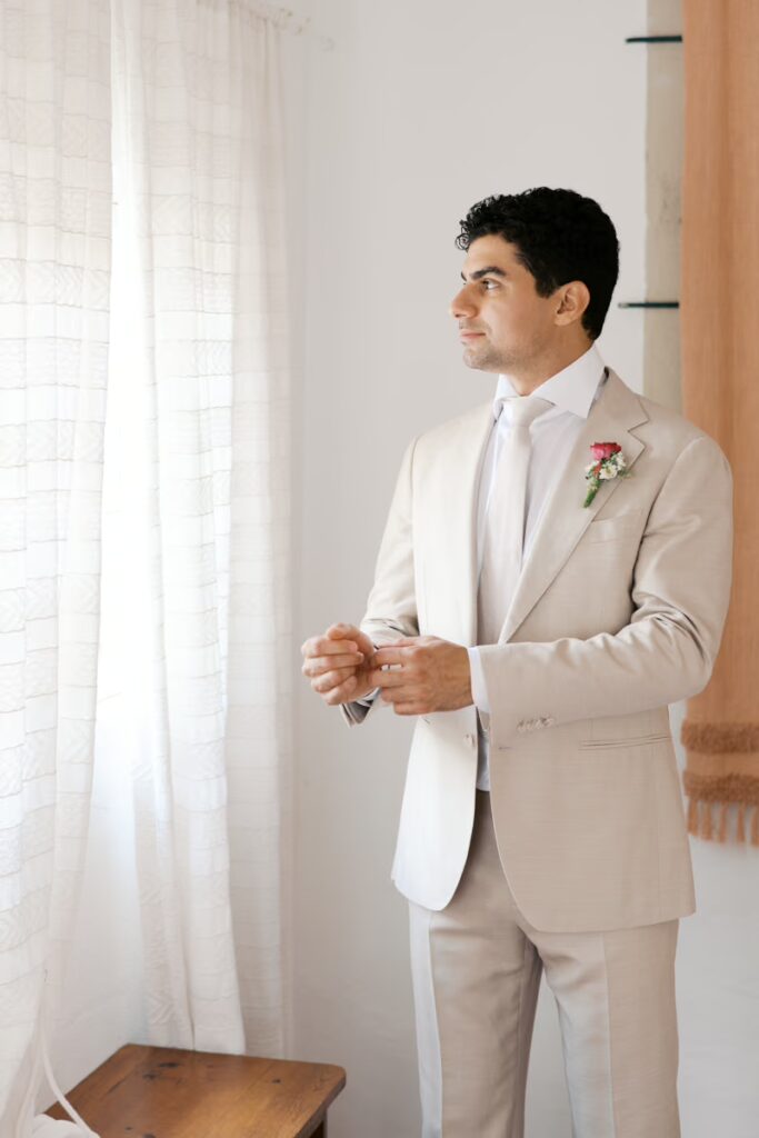 Retrato de perfil de um noivo em pé, ajustando as mangas de sua camisa social e as abotoaduras, olhando para a janela. Ele usa um terno bege claro areia bem cortado com uma gravata texturizada e uma boutonnière de flor rosa e cardo. A mesma cortina de linho texturizada é visível no fundo. Esta é uma foto de preparação no quarto da Quinta Nova do Hespanhol.