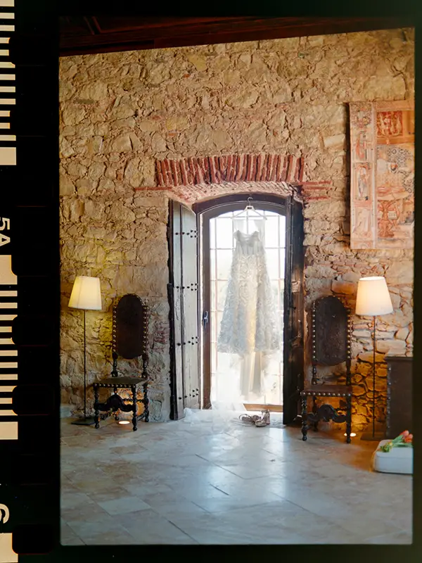 Wedding gown hanging in the natural light entry of a rustic room with ancient stone walls, at a historic estate in Portugal, Analogue Wedding Photography shot on 35mm film by Boris the Cat.